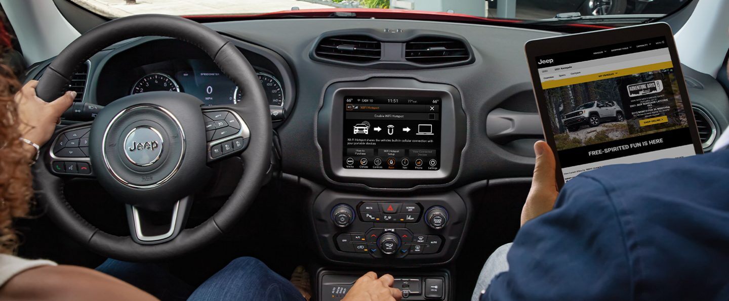 The Uconnect touchscreen on the 2021 Jeep Renegade displaying the Enable Wifi Hotspot screen and a passenger holding a tablet with jeep.com on-screen.
