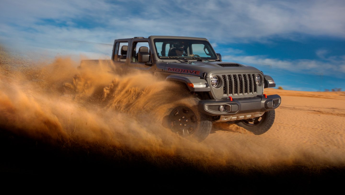 The 2022 Jeep Gladiator Mojave being driven off-road on sandy terrain with a large dust cloud obscuring its wheels.